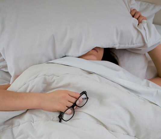 수면의 질이 건강을 좌우한다… 올바른 수면 습관 만들기의 중요성 person lying on bed while covering face with pillow and holding eyeglasses